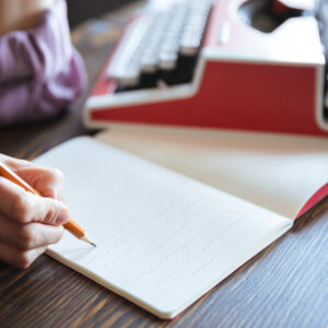 Retrato de uma mão feminina segurando um lápis sobre um caderno aberto, ao fundo uma máquina de escrever